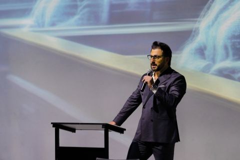 A person speaks into a microphone and leans on a podium in front of a digital screen