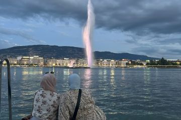 Two women in floral prints and head scarfs sitting on the waters edge at dusk with city lights and a small mountain in the background. 